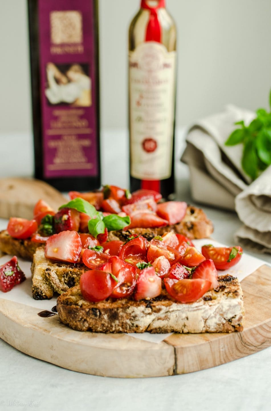 A side shot of strawberry tomato bruschetta on a cutting board with bottles of olive oil and balsamic vinegar in the background.