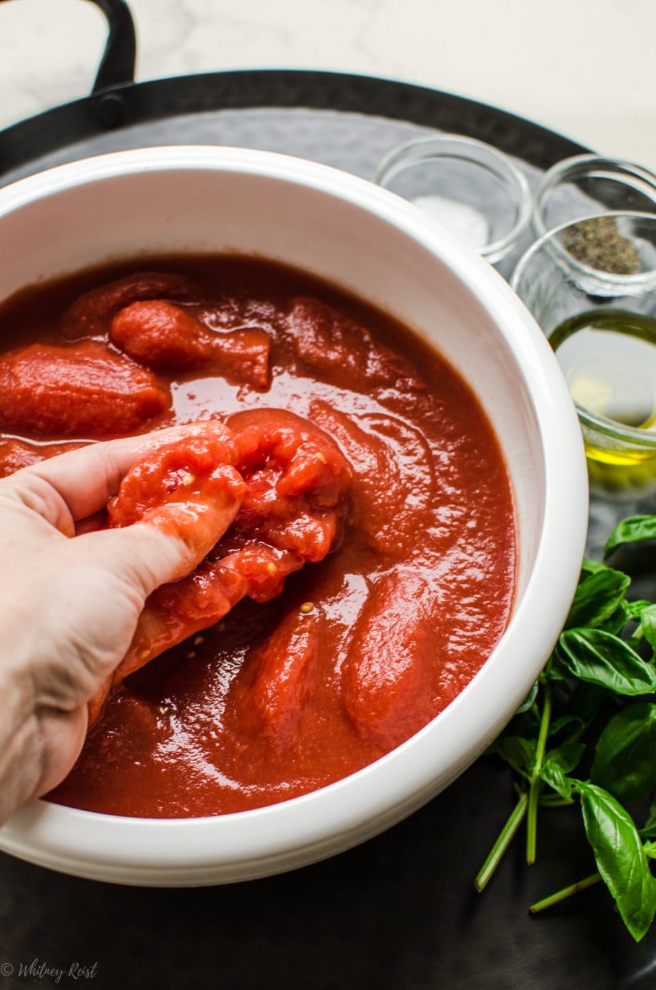 A hand squeezing a whole tomato into a white bowl to demonstrate making pizza sauce. 