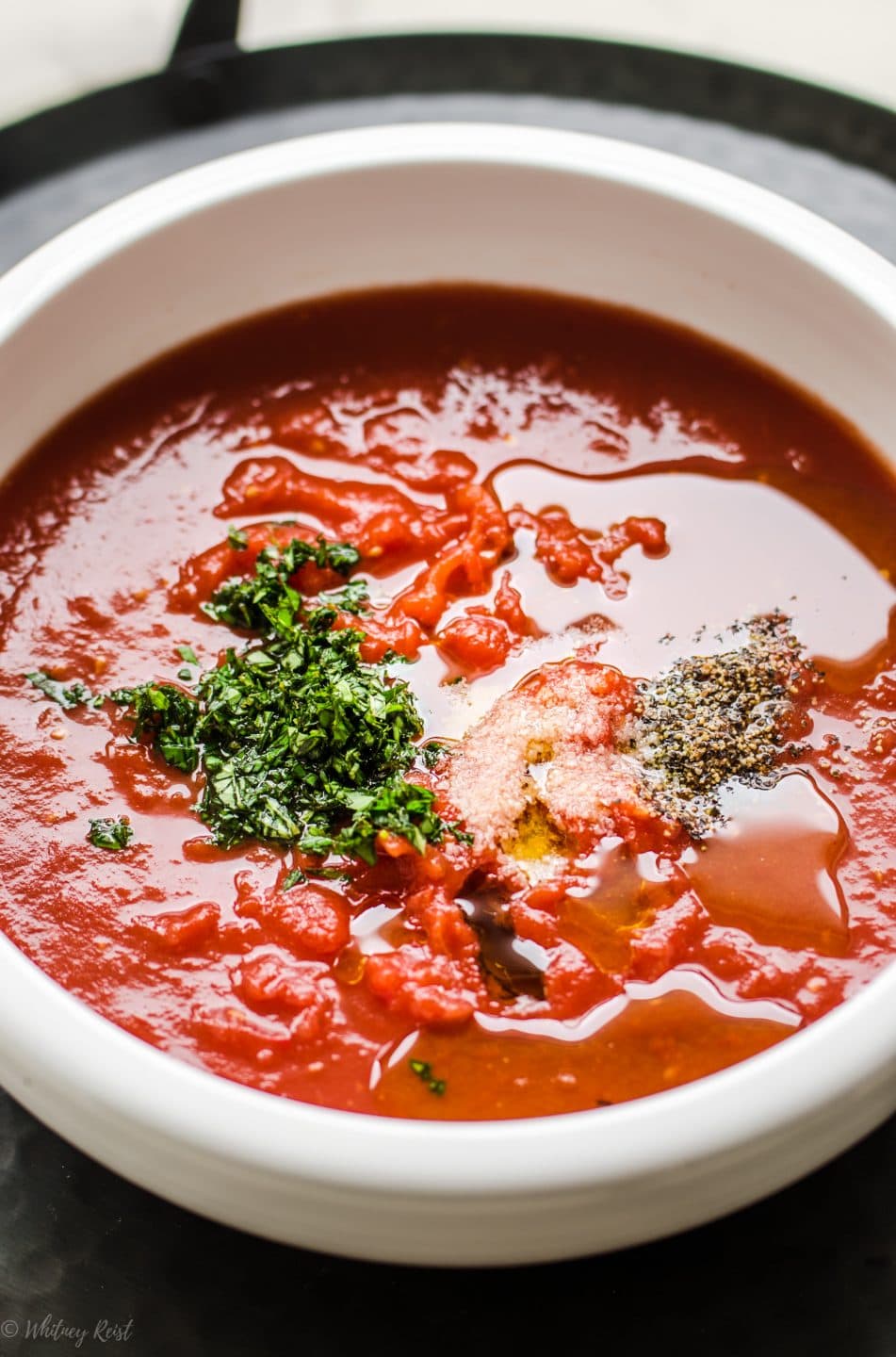 An overhead shot of a bowl of pizza sauce with fresh chopped basil, seasonings, and oil on the surface. 