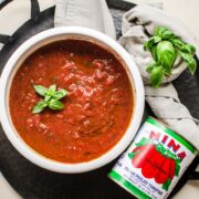 An overhead shot of a white bowl with tomato pizza sauce and can of San Marzano tomatoes on the side.