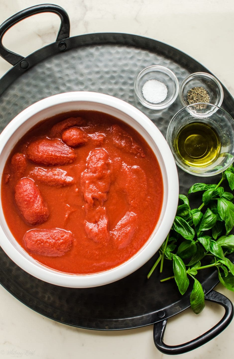 A white bowl with whole San Marzano tomatoes with olive oil, salt, pepper, and fresh basil on the side. 