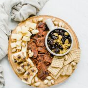 An overhead shot of a snacking board featuring beef jerky, crackers, dried fruit, cheese, and bananas topped with peanut butter and chocolate chips.