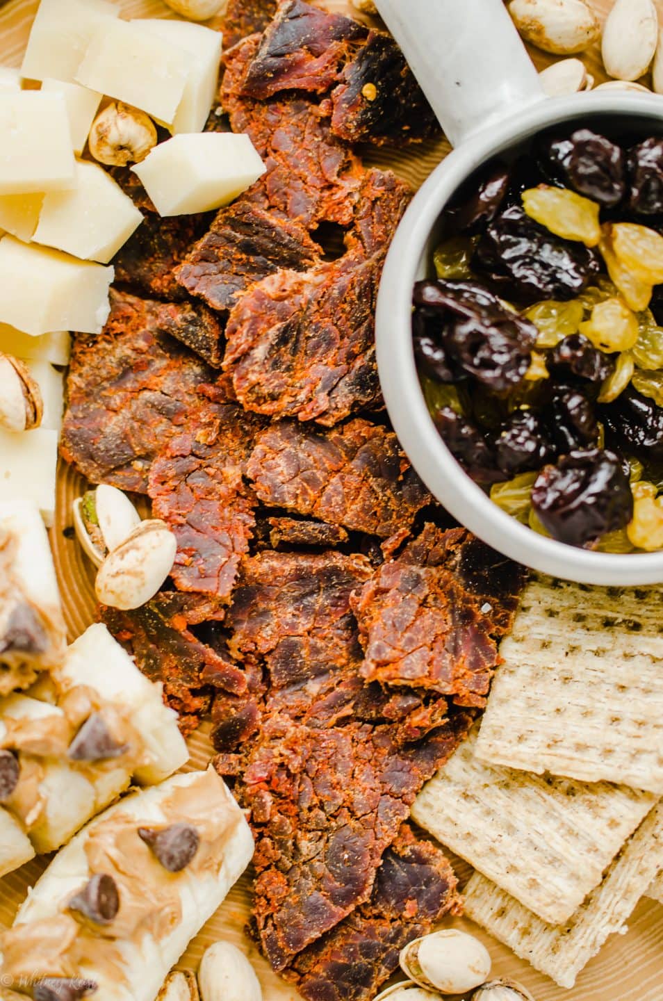 An overhead shot of a snacking board featuring beef jerky, crackers, dried fruit, cheese, and bananas topped with peanut butter and chocolate chips.