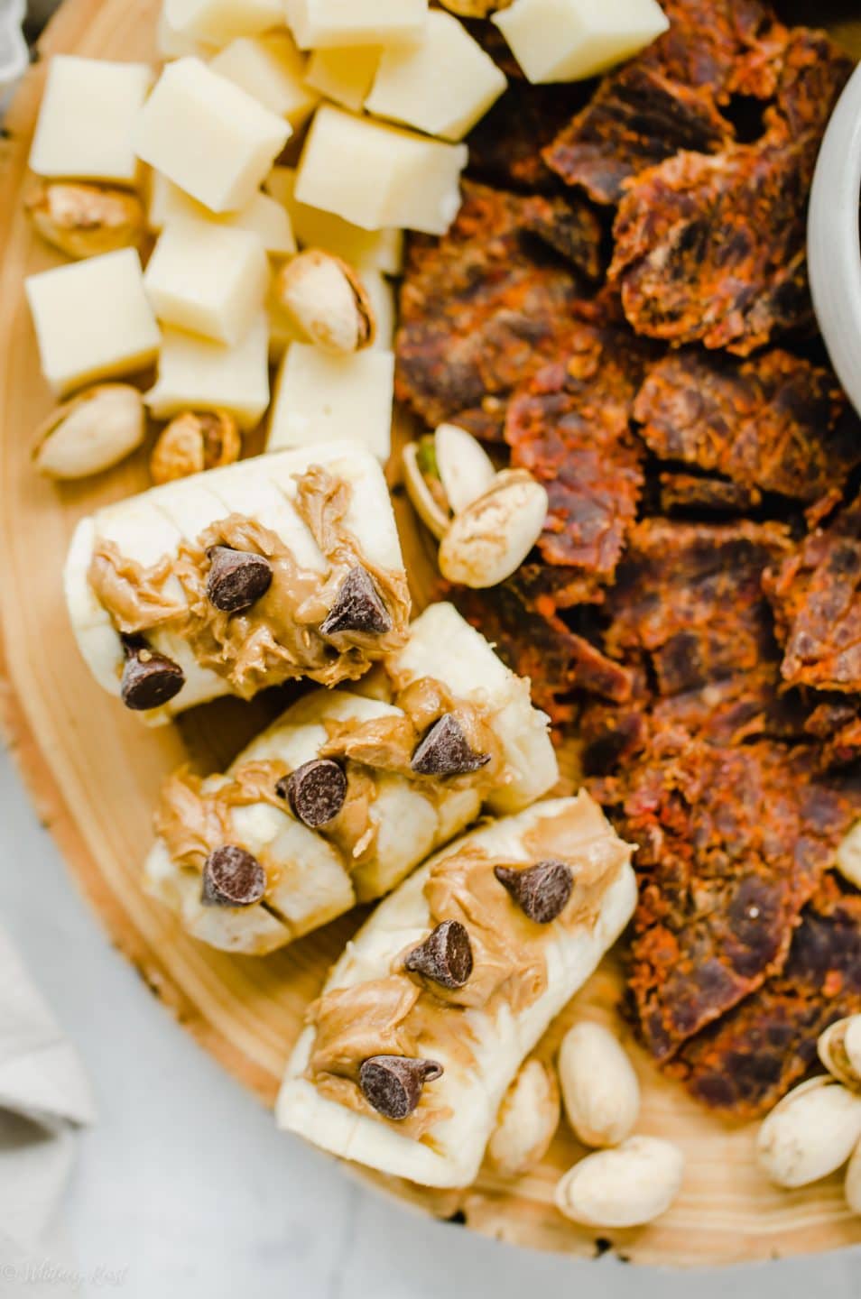 An overhead shot of a snacking board featuring beef jerky, crackers, dried fruit, cheese, and bananas topped with peanut butter and chocolate chips.