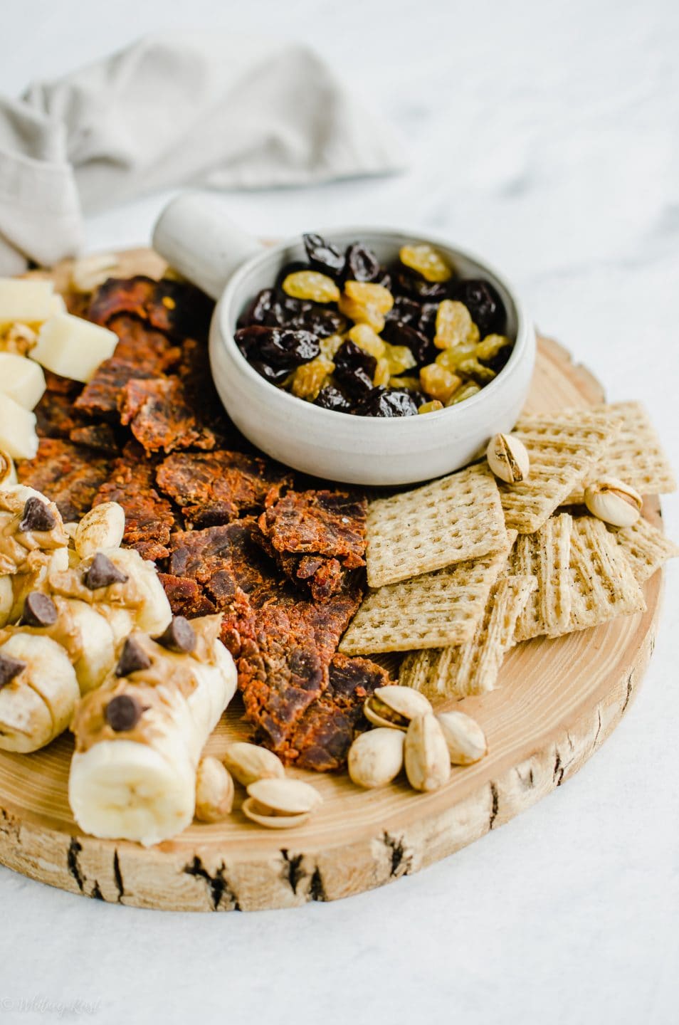 An overhead shot of a snacking board featuring beef jerky, crackers, dried fruit, cheese, and bananas topped with peanut butter and chocolate chips.