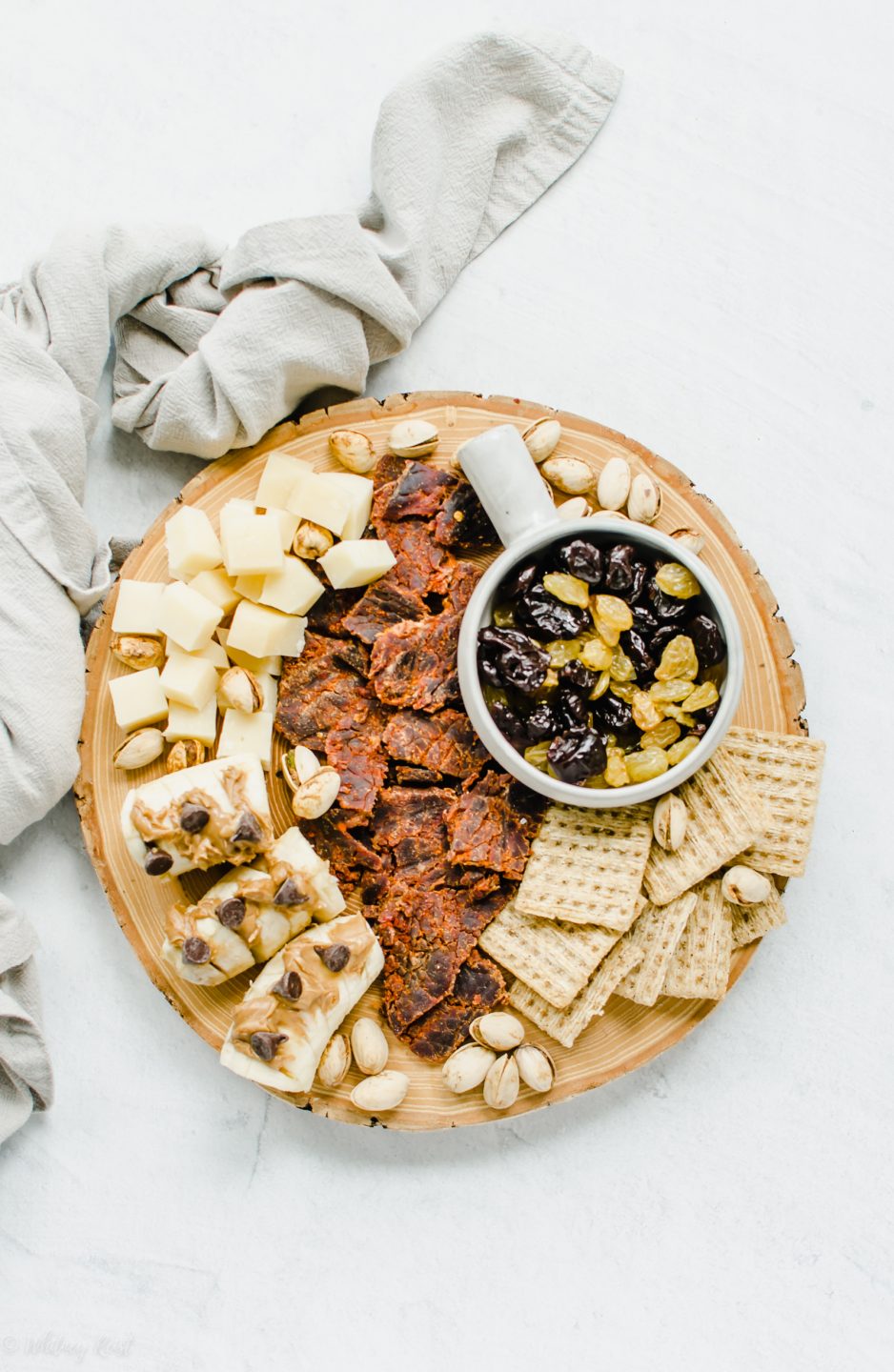 An overhead shot of a snacking board featuring beef jerky, crackers, dried fruit, cheese, and bananas topped with peanut butter and chocolate chips. 