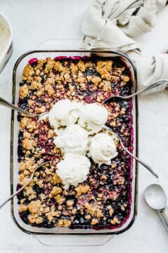 A rectangular baking dish with mixed berry crumble inside and scoops of ice cream on top with spoons on the side.