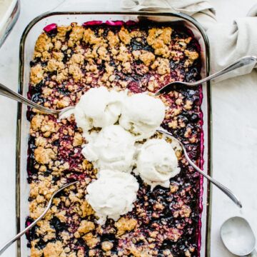 A rectangular baking dish with mixed berry crumble inside and scoops of ice cream on top with spoons on the side.