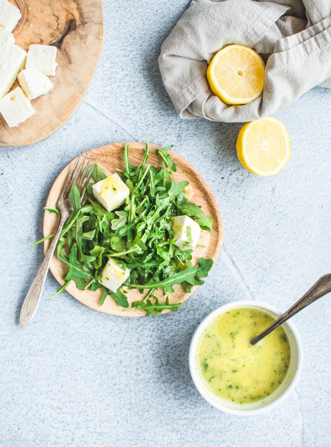 A wooden plate with arugula feta salad and a white bowl of mint vinaigrette on a blue background. 