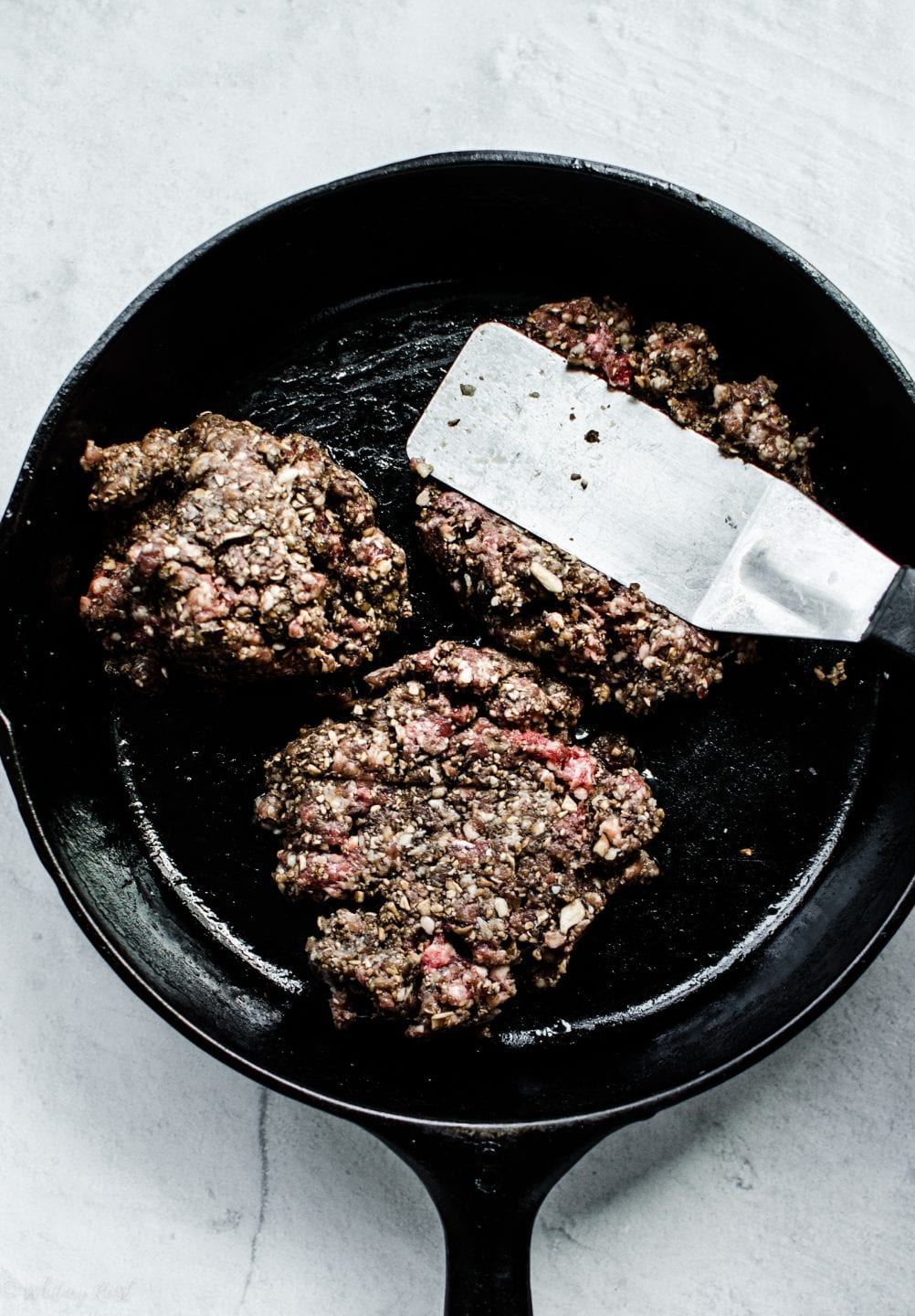 3 mounds of mushroom ground beef blend in a cast iron skillet with a spatula smashing one of them. 
