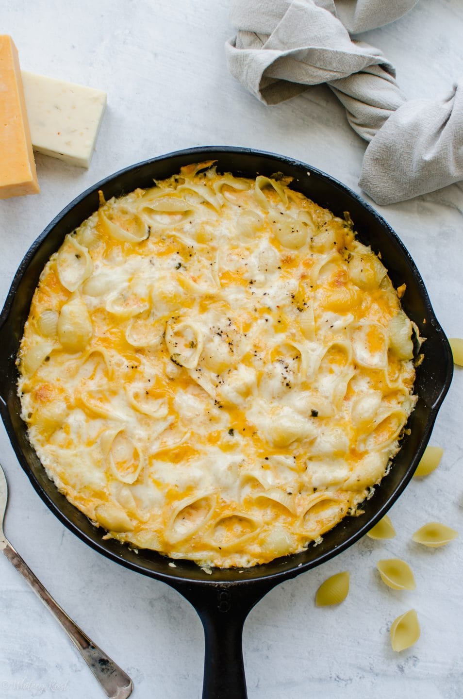 An overhead shot of a cast iron skillet of baked mac and cheese with dried pasta and cubes of cheese around it.