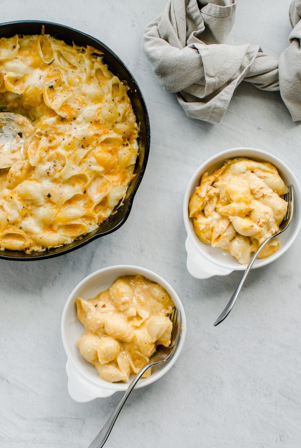 Two white dishes of mac and cheese next to a larger skillet of mac and cheese on a white stone backdrop.