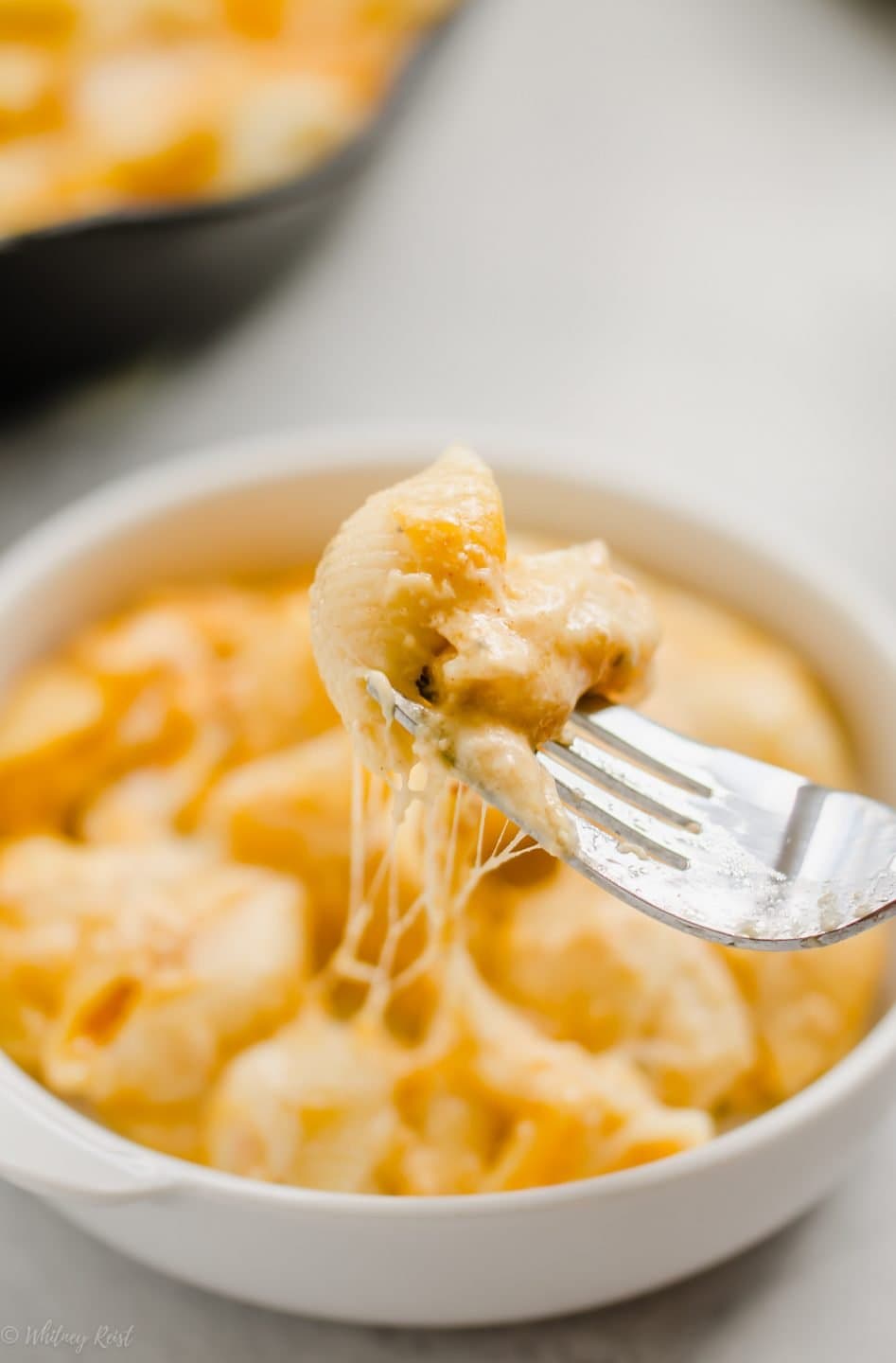 A close up shot of a fork with a bite of mac and cheese with strings of cheese coming from the dish below.