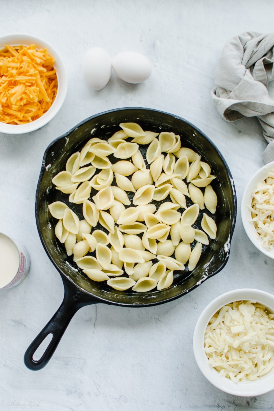 A cast iron skillet with noodle shells inside and bowls of mac and cheese ingredients surrounding the skillet.