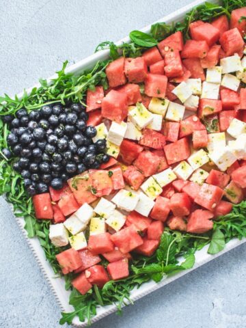 A white platter with a red, white, and blue watermelon flag salad on top on a blue stone background.
