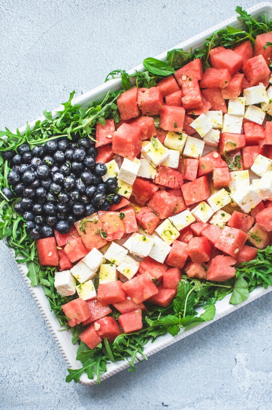 A white platter with a red, white, and blue watermelon flag salad on top on a blue stone background.