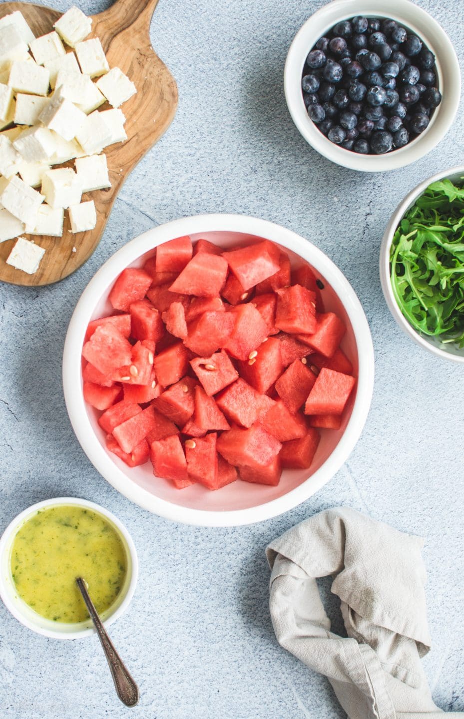 A white bowl filled with watermelon cubes surrounded by smaller white bowls with feta cheese, blueberries, arugula, and mint dressing.