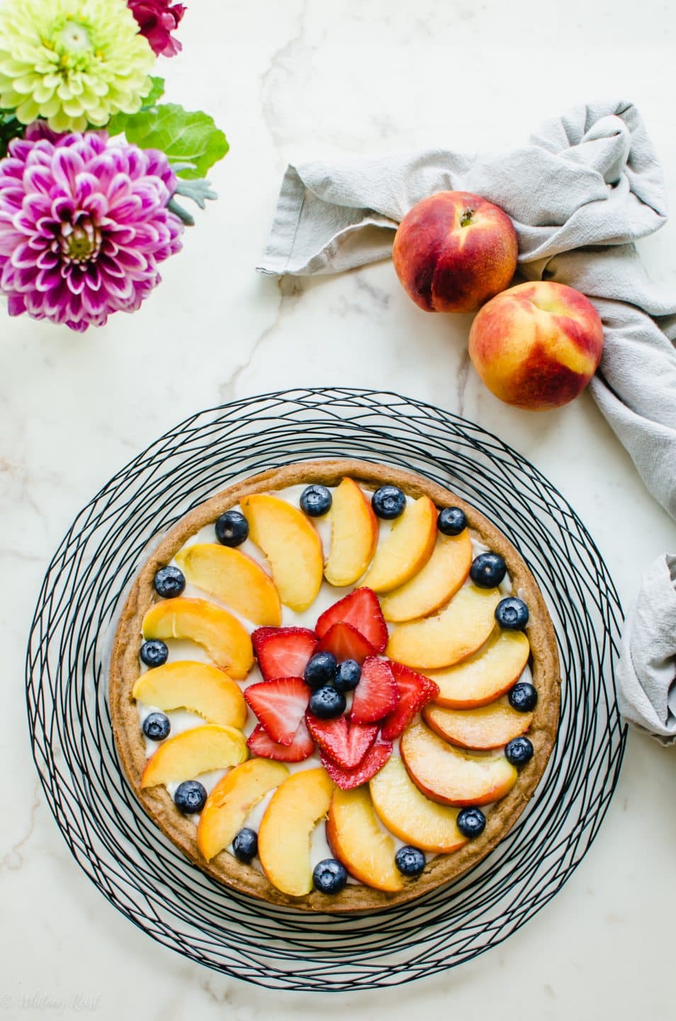 A peach fruit pizza on a wire charger with whole peaches and a vase of flowers on the side.