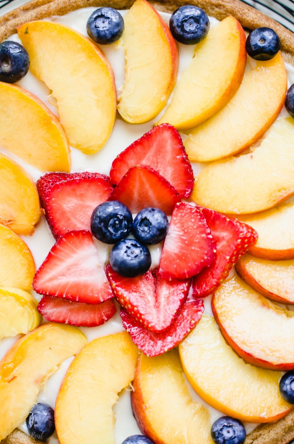 A close up shot of the middle of a decorated fruit pizza with peaches, strawberries, and blueberries.