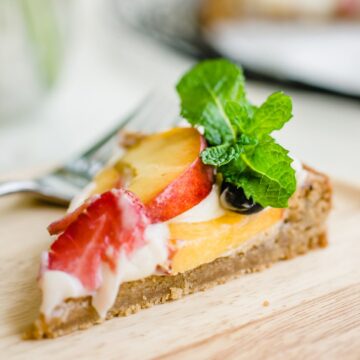 A side angle shot of a slice of peach fruit pizza on a wooden plate garnished with a sprig of mint.