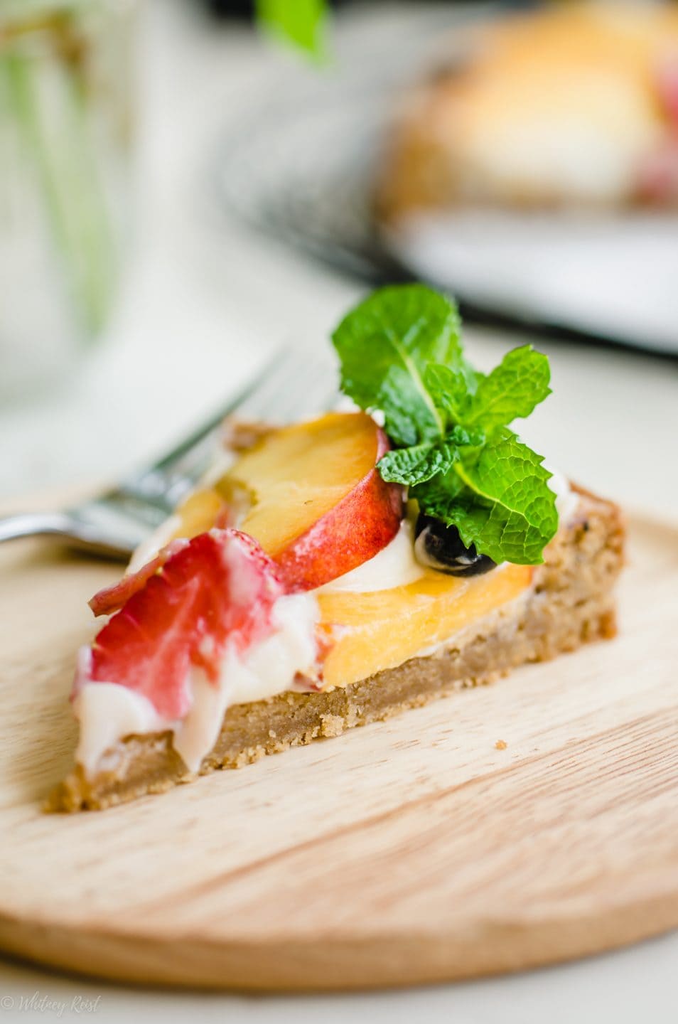 A side angle shot of a slice of peach fruit pizza on a wooden plate garnished with a sprig of mint.