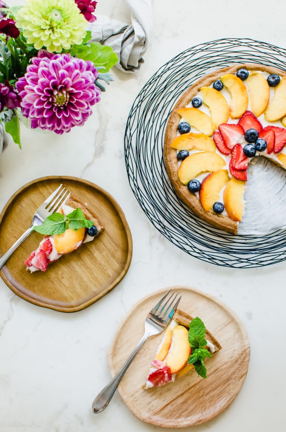 An overhead shot of two wooden plates of fruit pizza with forks and a vase of flowers.