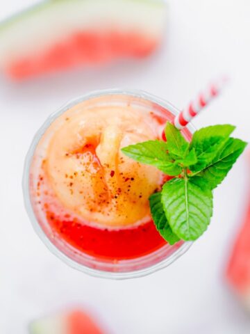 An overhead shot of the top of a glass filled with watermelon juice and mango sorbet with a red striped straw and sprig of mint.