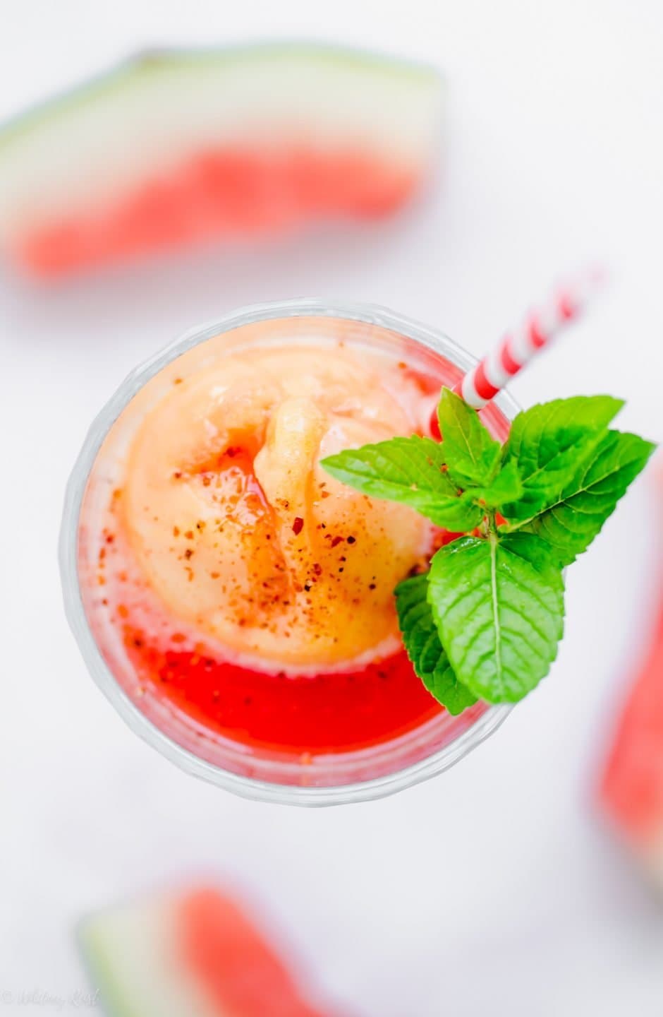 An overhead shot of the top of a glass filled with watermelon juice and mango sorbet with a red striped straw and sprig of mint.