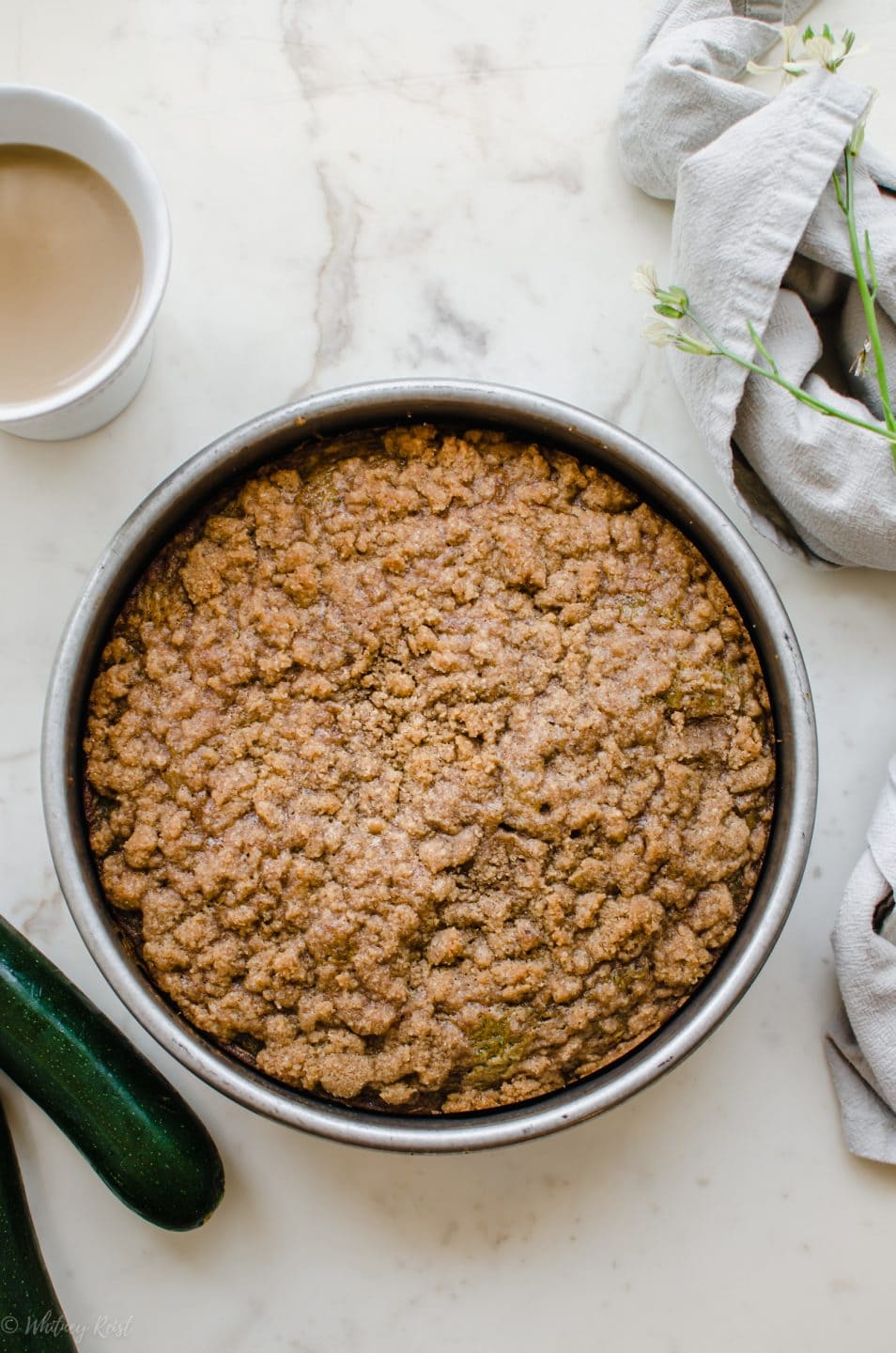 A zucchini coffee cake in a cake pan with a cup of coffee on the side.