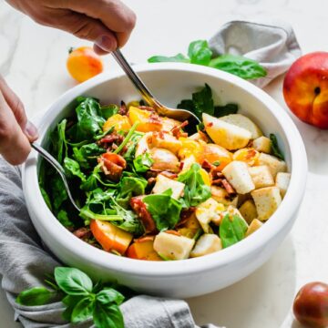 A demonstration of hands tossing a BLT Panzanella Salad in a white ceramic serving bowl.