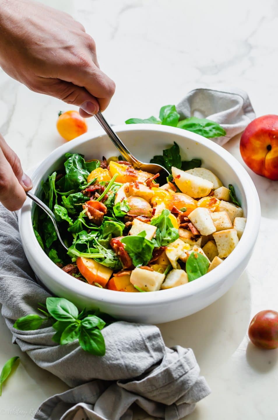 A demonstration of hands tossing a BLT Panzanella Salad in a white ceramic serving bowl.