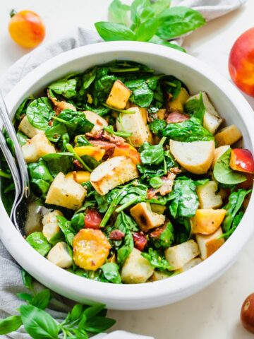 A white bowl filled with a tossed BLT Panzanella Salad with silver serving utensils on the side.