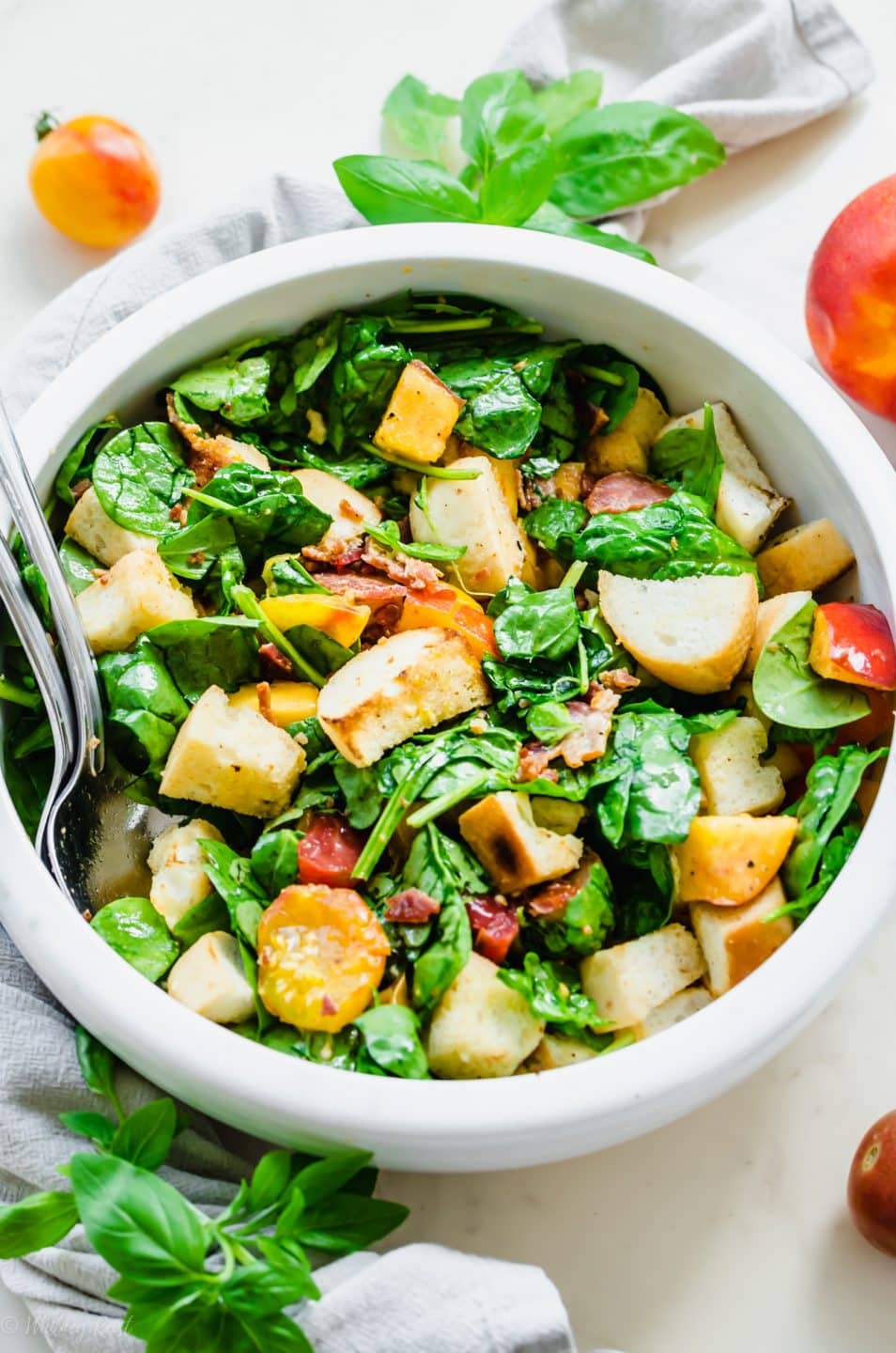 A white bowl filled with a tossed BLT Panzanella Salad with silver serving utensils on the side.