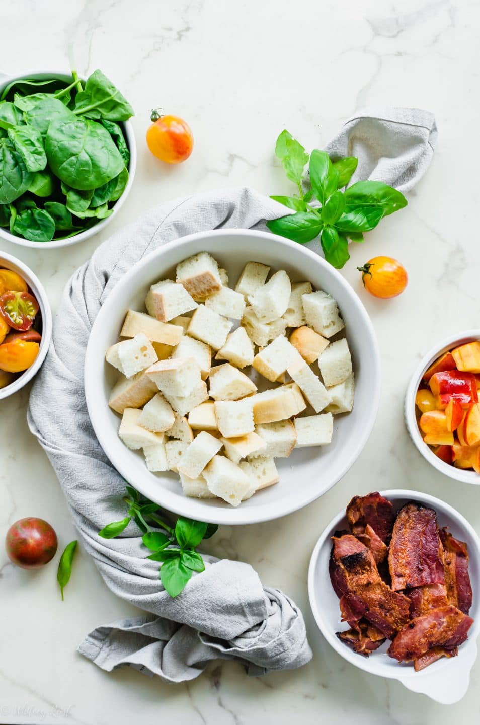 White bowls of ingredients for making BLT Panzanella Salad on a white marble counter top.