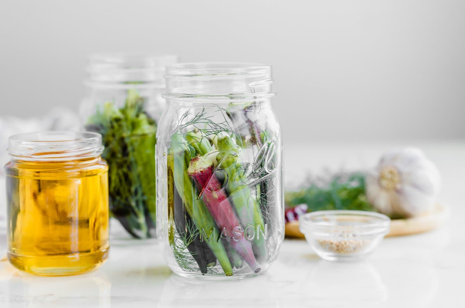 A canning jar with fresh okra with apple cider vinegar, garlic, and dill on the side.