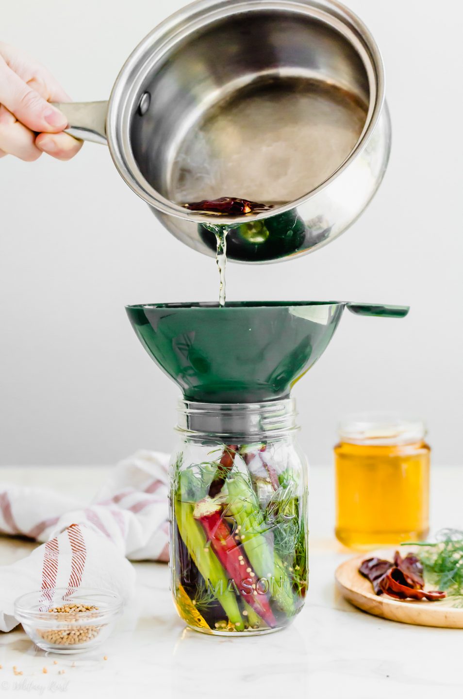 A hand pouring pickling liquid into a jar of pickled okra.