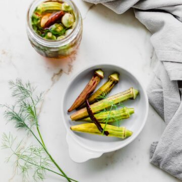 An overhead shot of a white dish with pickled okra and a jar of pickled okra on the side.