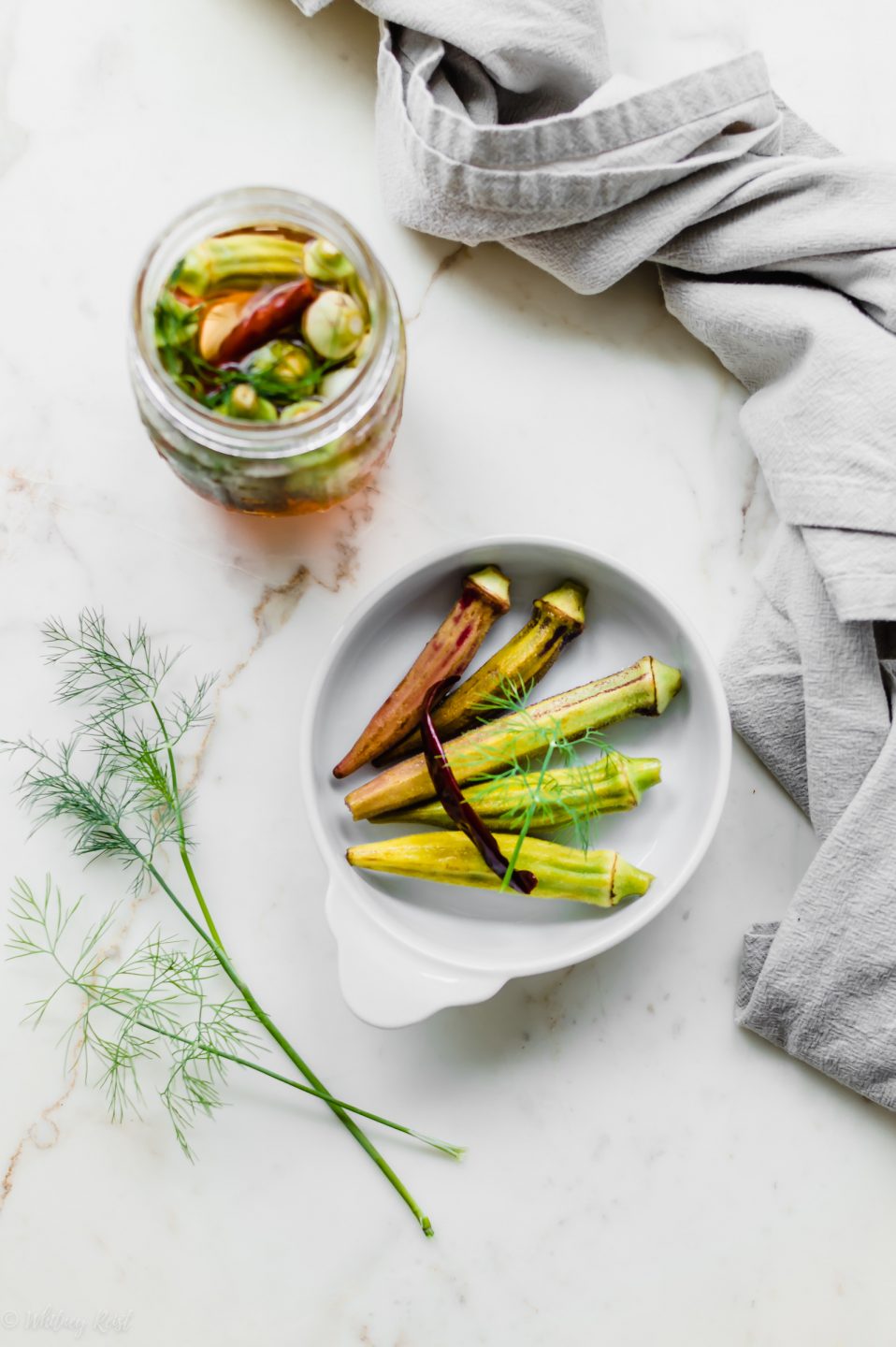 An overhead shot of a white dish with pickled okra and a jar of pickled okra on the side.