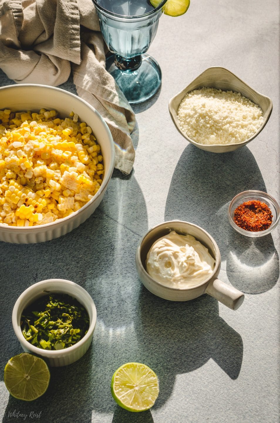 All of the ingredients for elote-style corn in prep bowls on a blue concrete background.