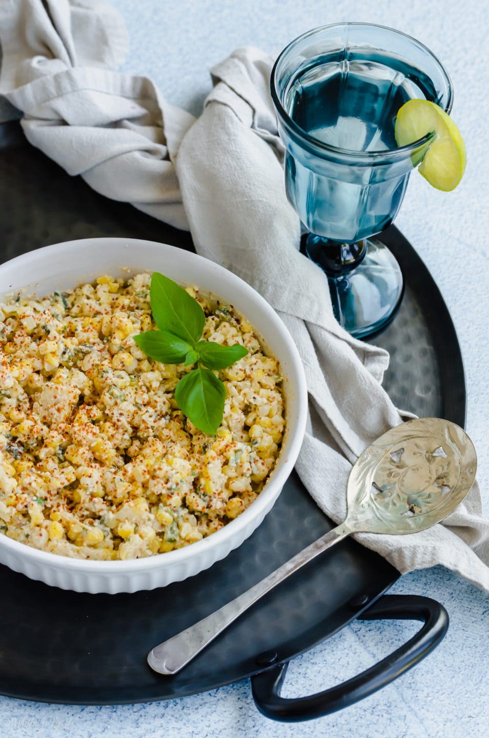 A bowl of elote-style corn on a black platter with a glass of blue water.