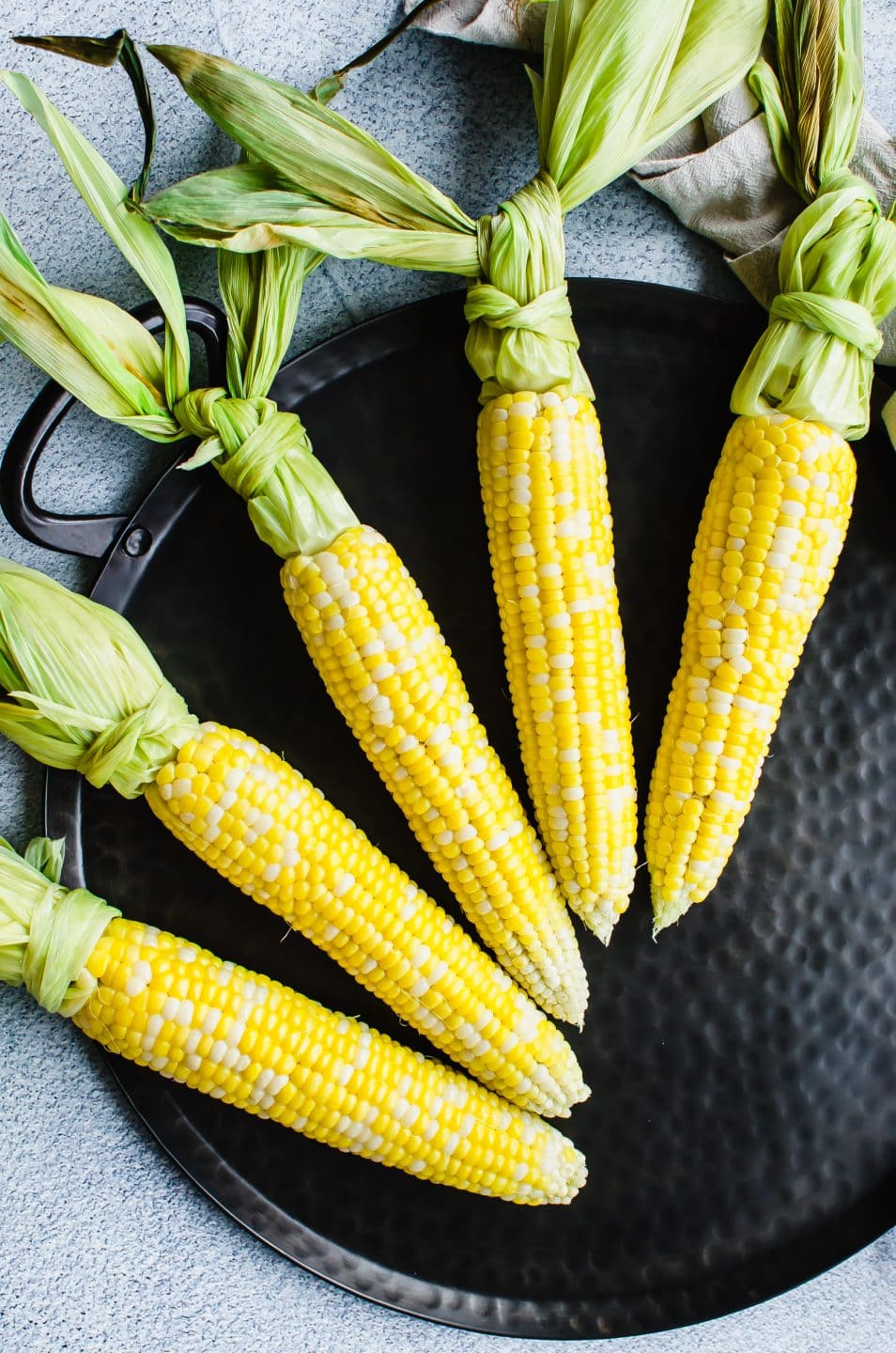 Raw corn on the cob on a black iron platter with the husks tied in knots.