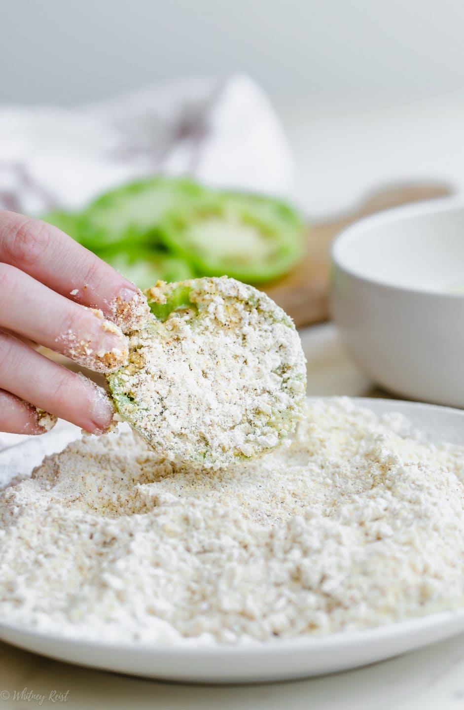 A hand dipping a sliced green tomato in a cornmeal bread crumb mixture.