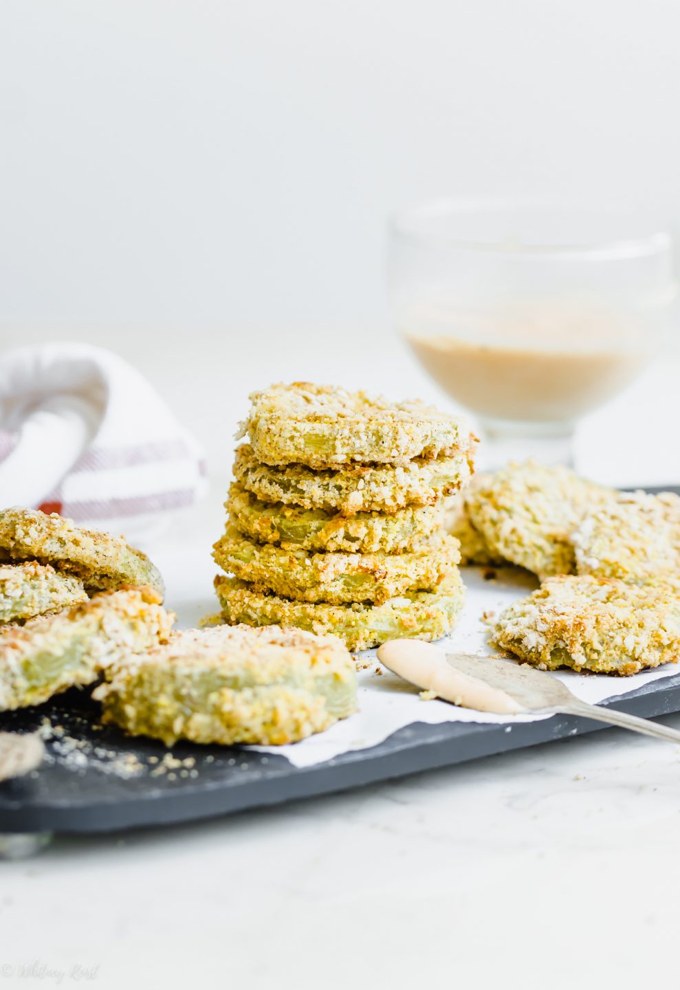 Oven-fried green tomatoes stacked on a slate platter with a bowl of hot sauce aioli on the side.