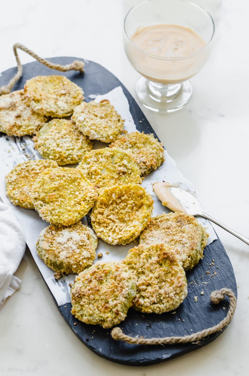 A slate platter lined with parchment paper and topped with fried green tomatoes and a bowl of hot sauce aioli on the side.