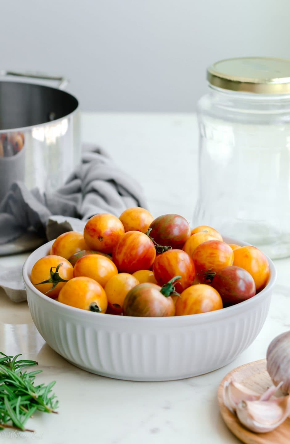 A straight-on shot of the jar, tomatoes, pot, rosemary, and garlic cloves needed for a pickled tomatoes recipe.