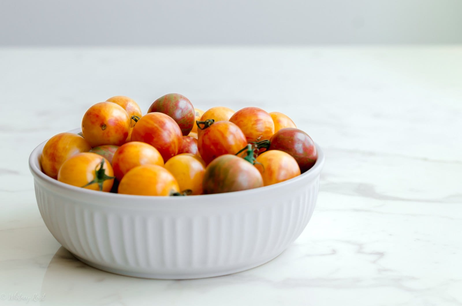 A side angle of tomatoes in a white bowl on a white marble countertop.