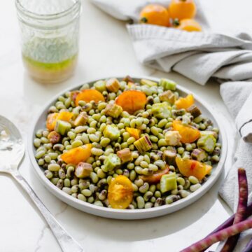 A gray plate with purple hull pea salad and a jar of vinaigrette, tomatoes, and peas in the shell on the side.