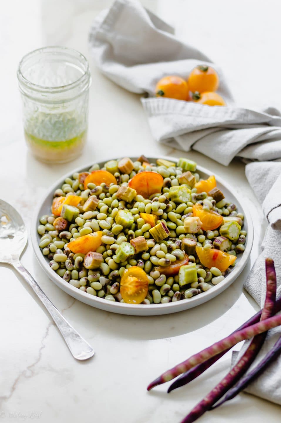 A gray plate with purple hull pea salad and a jar of vinaigrette, tomatoes, and peas in the shell on the side.