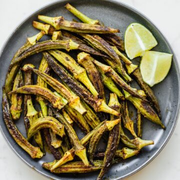 An overhead shot of a gray ceramic plate with roasted okra and lime wedges.