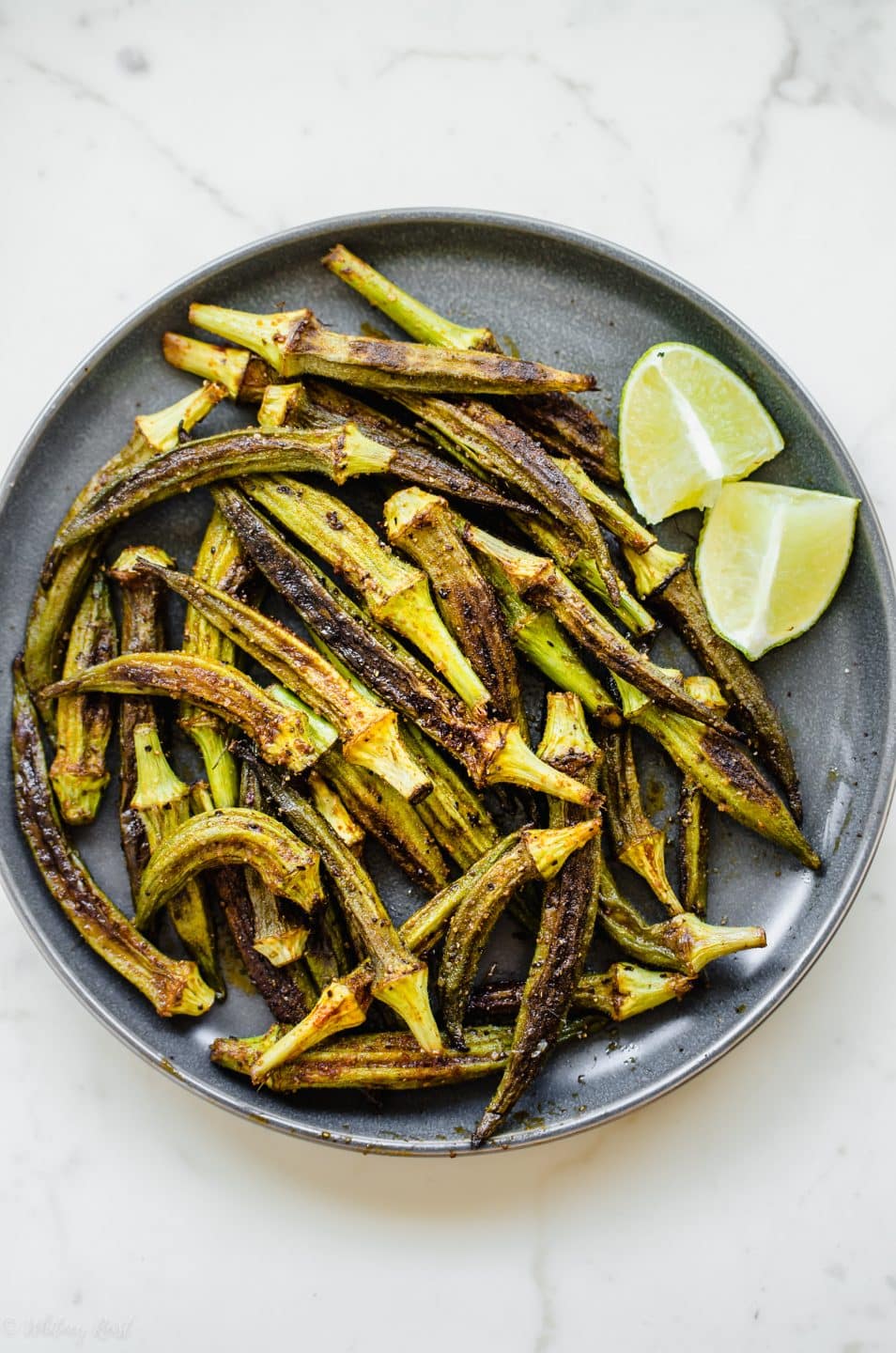 An overhead shot of a gray ceramic plate with roasted okra and lime wedges.